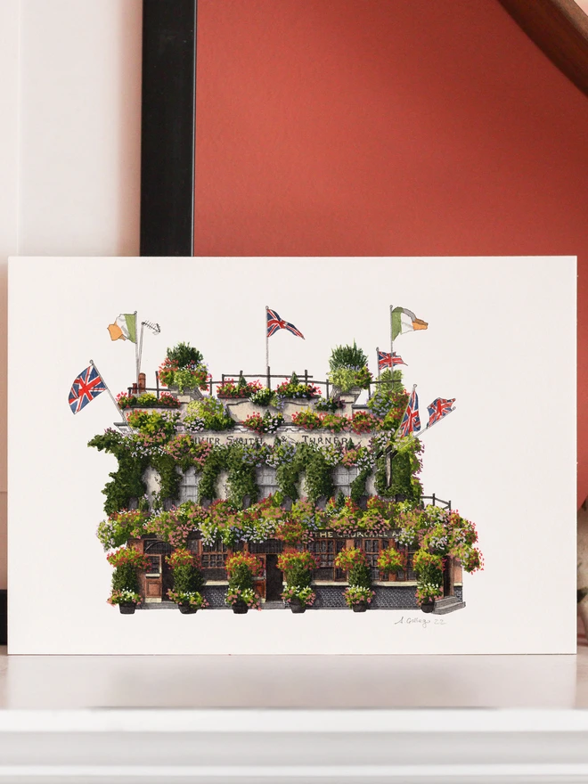 An architectural illustration of The Churchill Arms pub on a print, displayed on a white shelf alongside cat and elephant figurines against a pink wall.