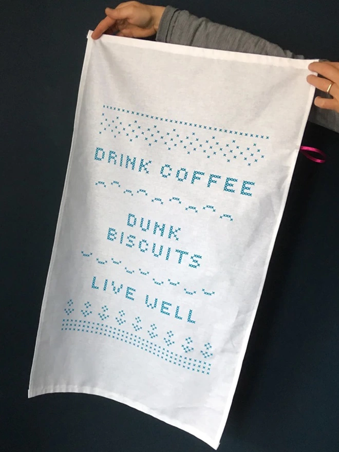 A White cotton tea towel with cross stitch lettering," Drink Coffee, Dunk biscuits, Live Well' being held in front of a dark background