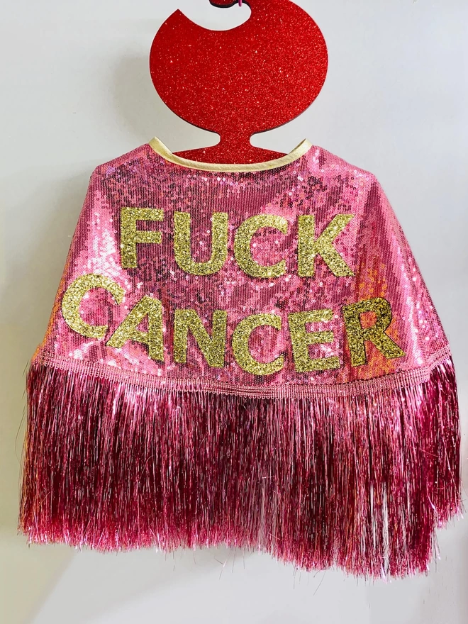 A cape hangs on a hook. The words 'FUCK CANCER' are across the back in gold glitter against pink sequins. The cape has a gold neck tie and pink tinsel.