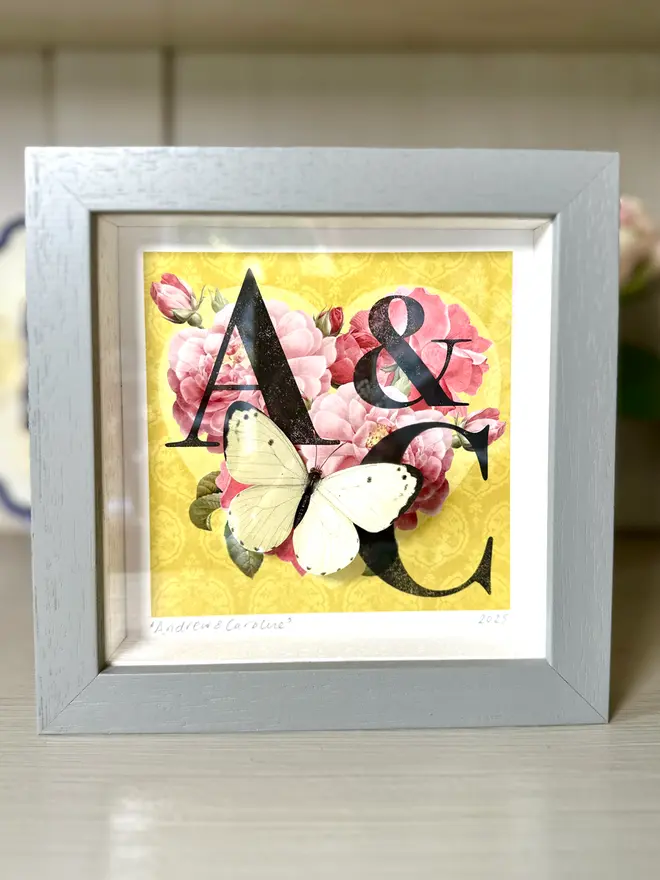 A hand made Golden wedding anniversary card featuring a heart made of deep pink roses on a golden background with letters and initials of the couple. A white paper cut three dimensional butterfly sits on the heart. The hand made card is in a light grey wooden frame showing the gift option of this keepsake card.
