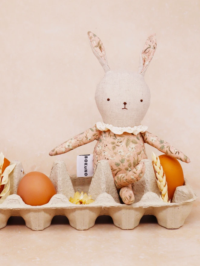 Handmade linen bunny doll in pink floral outfit and frilly lace colour, sitting in an egg carton with eggs.