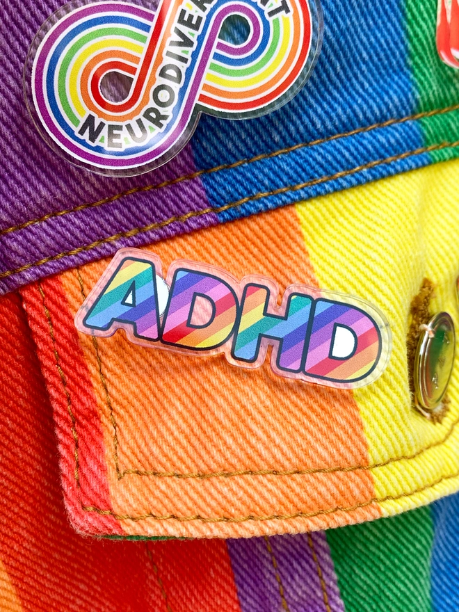 image shows an acrylic badge in the shape of the letters ADHD. the letters are filled with diagonal rainbow stripes. the badge is pinned to the pocket of a rainbow striped denim jacket.