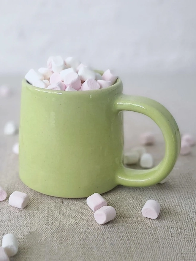 Ceramic Standard Mug, a colourful ceramic mug sitting on a plain surface against a grey backdrop. It is accompanied by marshmallows. 