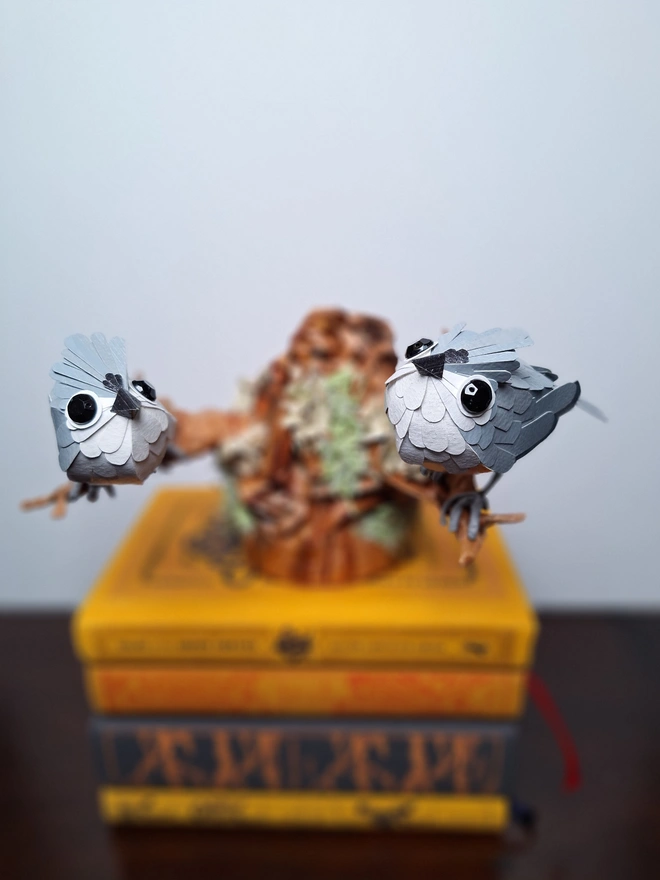 A handmade paper sculpture depicting two tufted titmice birds perched on a tree stump. The sculpture is placed on top of a pile of books. Focus is specifically on the two birds, showing their individual feathers and markings.