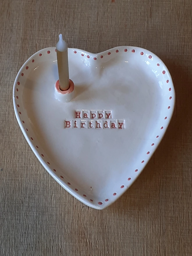 Handmade white heart-shaped ceramic Happy Birthday dish with red lettering, red dotted edging and a candle in the built-in holder.
