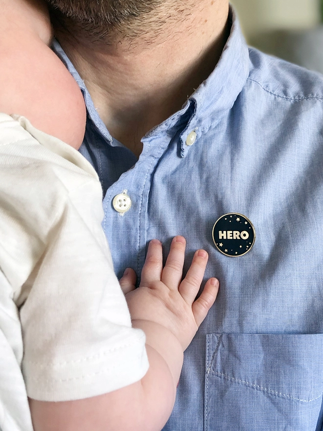 A navy blue and gold enamel pin badge with a starry design and the word "Hero" is pinned to a man's blue shirt while he is holding a baby.