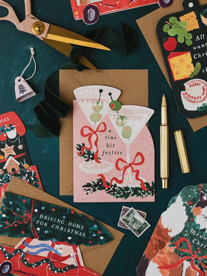 Tini Bit Festive Christmas card on a dark green background with the Driving Home For Christmas and Cheeseboard cards, gold scissors, green ribbon and a gold pen