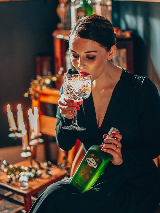 Holly drinking from a vintage glass and holding a Holly's Gin bottle