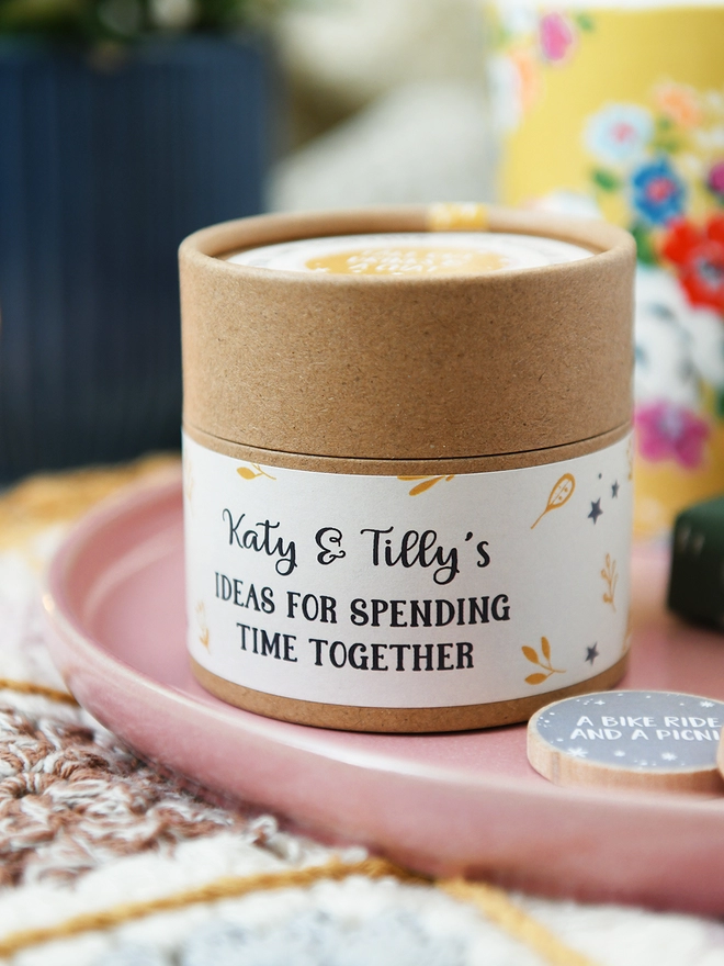 A cardboard jar with a floral label on the side that reads Ideas for spending time together stands on a pink plate.