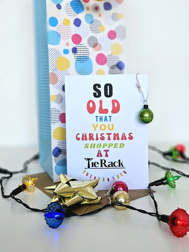 A photograph of the front of a humorous A6 greeting card illustrated using brightly coloured text and the retro shop logo. The text says so old you Christmas shopped at Tie Rack  with a string of fairy lights