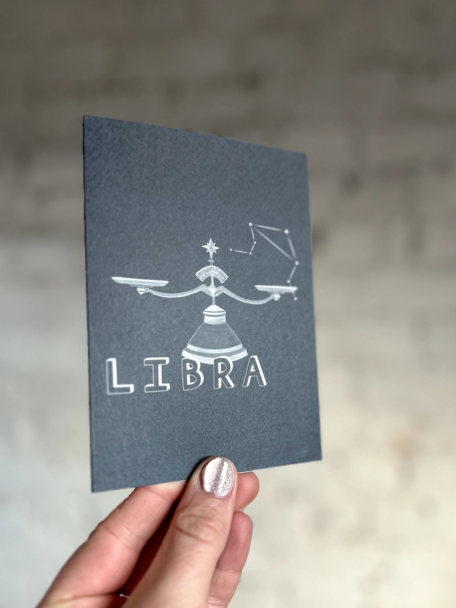  a greetings card featuring a greyscale illustration of a zodiac style set of scales with the text “LIBRA”