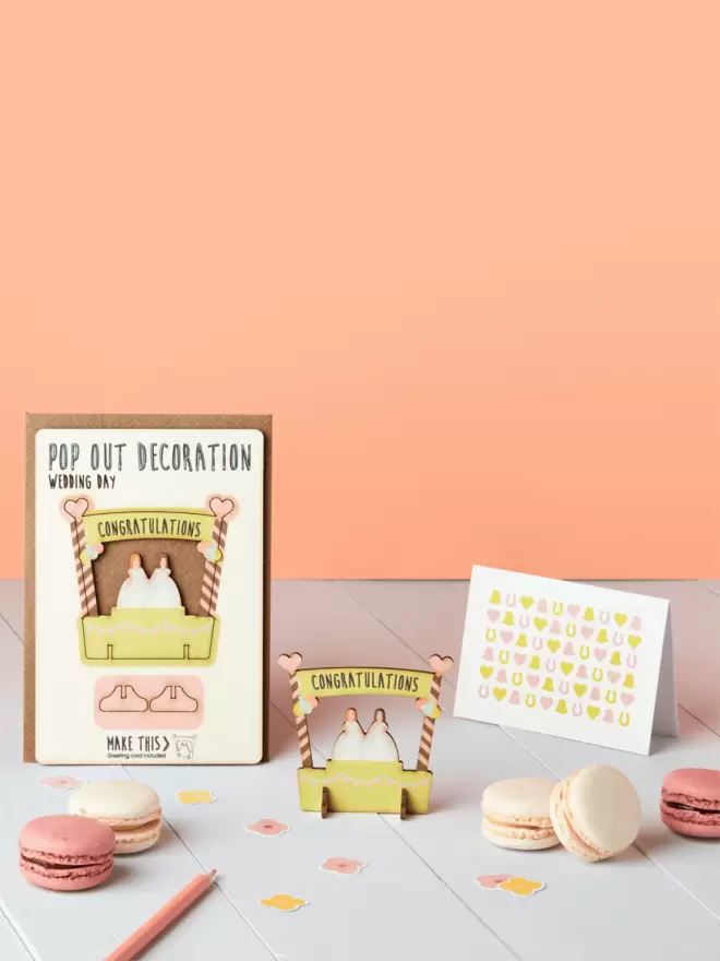 Pop out two brides wedding decoration and confetti pattern wedding card and brown kraft envelope on top of a wooden desk in front of a peach coloured background