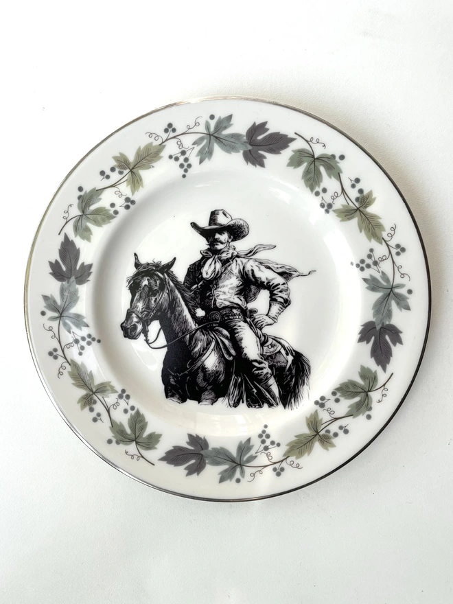 vintage plate hanging on a white wall, it has a decorative border of grey and blue leaves, with a printed vintage illustration of a cowboy in the middle 