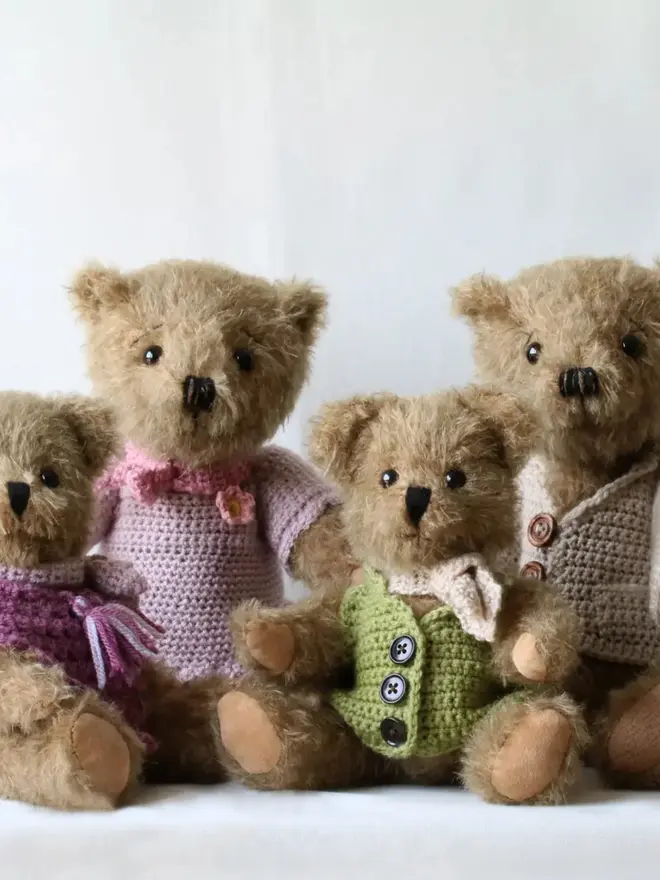 Handmade brown teddy bear wearing a green cardigan and white scarf, sitting with other teddy bears against a plain background.