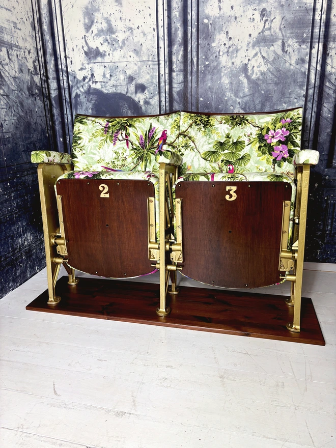 set of two vintage cinema seats upholstered in a purple and green macaws velvet.  Seats are sat on a white wooden floor with a marbled blue wall behind them