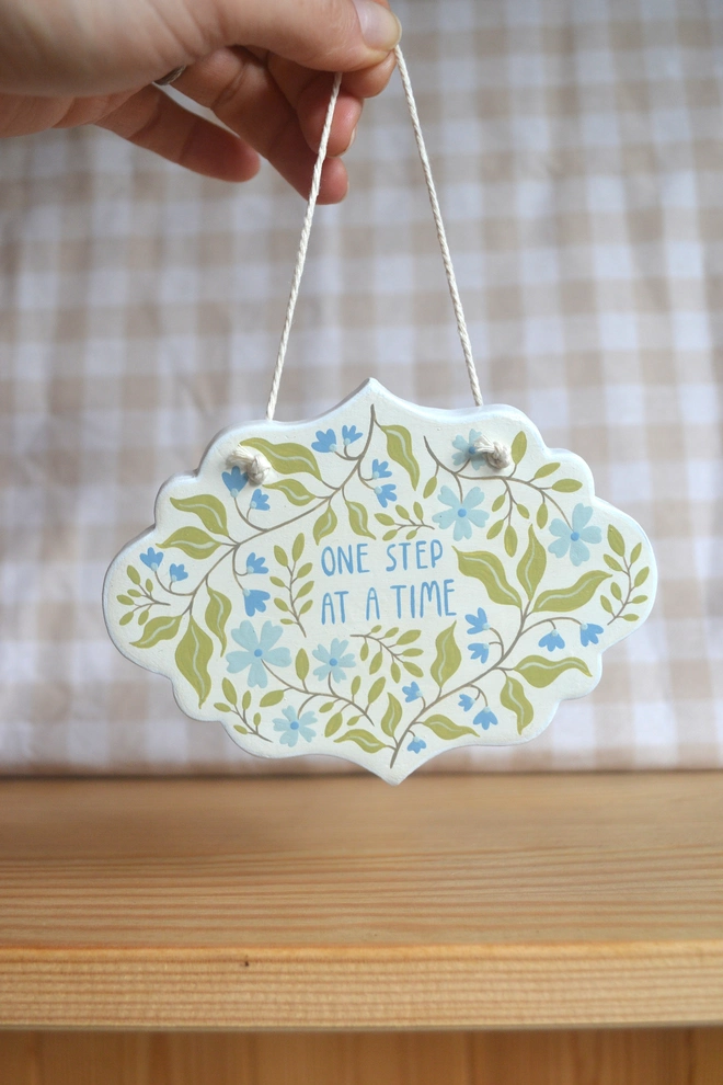 hanging clay sign with blue and green floral design reading 'one step at a time' in front of gingham background