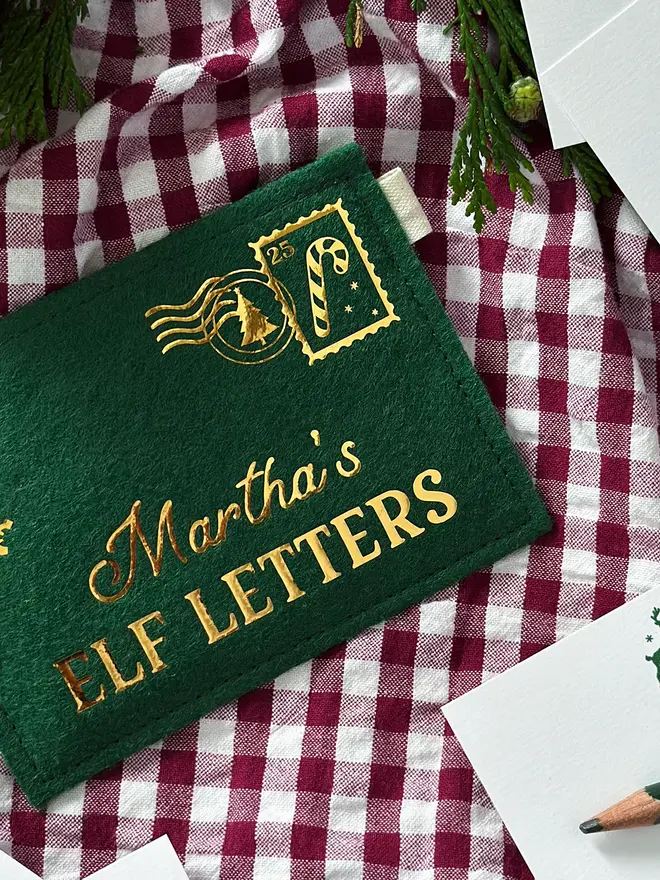A green felt reusable envelope with the words "Martha's Elf Letters" in golden writing and a candy cane stamp in the top right corner, lays on red gingham fabric.