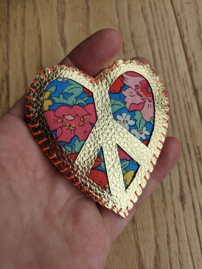 Gold faux leather and Liberty floral fabric Peace sign heart brooch being held by a hand