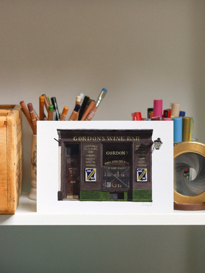 A watercolor print of Gordon's Wine Bar displayed on a shelf alongside various art supplies