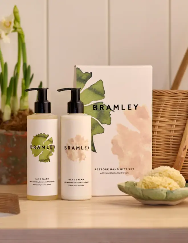 two bottles of product with white labels and black caps, next to a white box with green and pink floral design, on a beige table with plants and a wicker basket in the background
