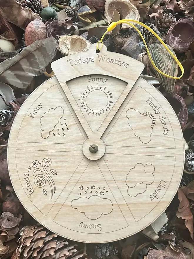 Extra large wooden weather wheel with engraved weather symbols for sunny, cloudy, rainy, windy, snowy, and partly cloudy, shown resting among pinecones and natural materials.