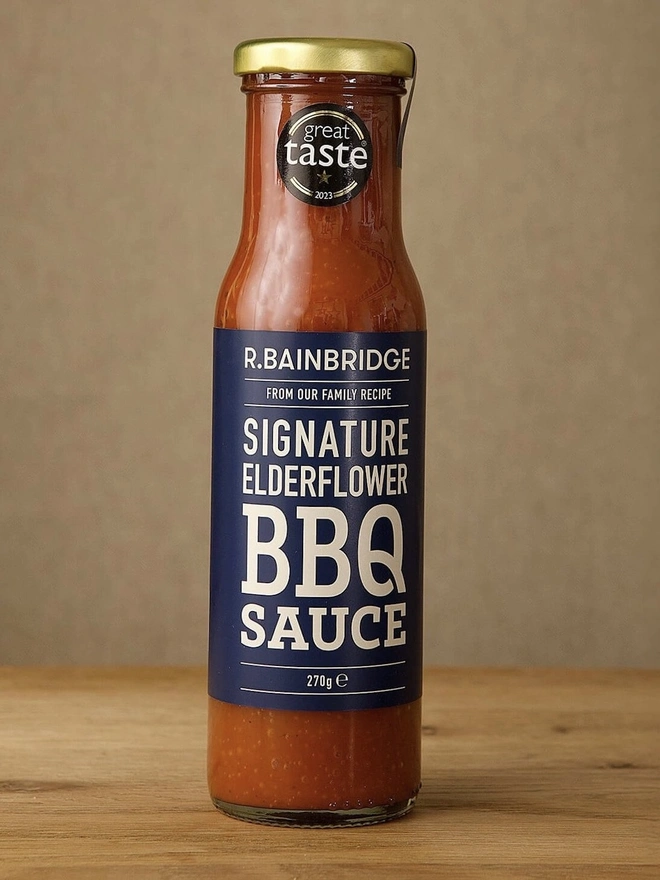 Elderflower BBQ Sauce, a jar of BBQ sauce sitting on a wooden surface in front of a plain backdrop. 