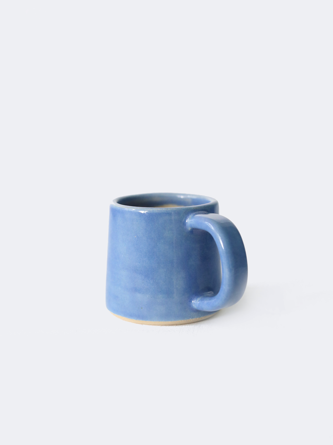 A handmade ceramic espresso mug in pacific blue glaze, with a chunky loop handle, photographed against a plain light grey background