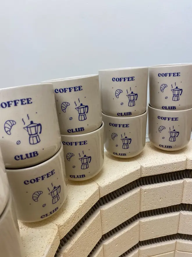 'Coffee Club' Handmade Ceramic Coffee Cup, a group of coffee cups stood against a white backdrop with blue designs. 