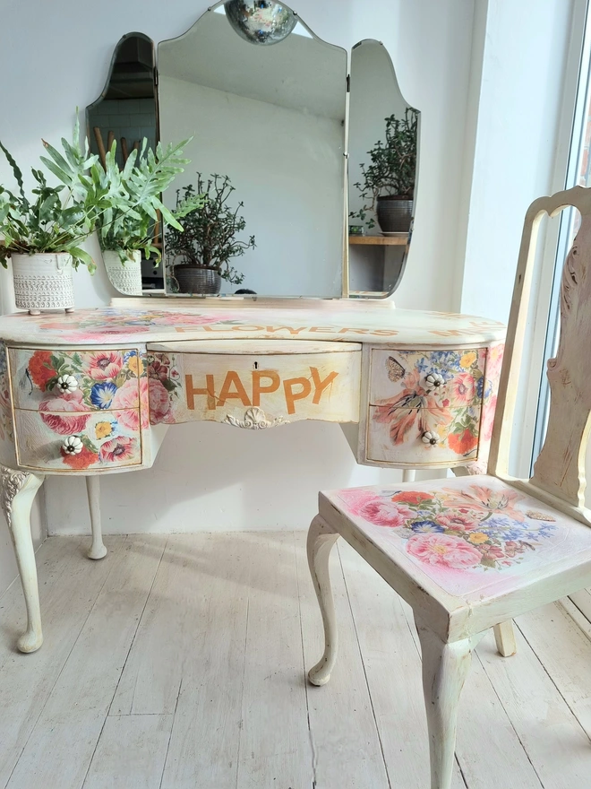 An antique kidney-shaped dressing table with chair and a trio of mirrors. Upcycled in cream and pale pink with decoupage florals in oranges and pinks. Text across the table read ‘flowers make me happy’ in the orange of the undercoat.