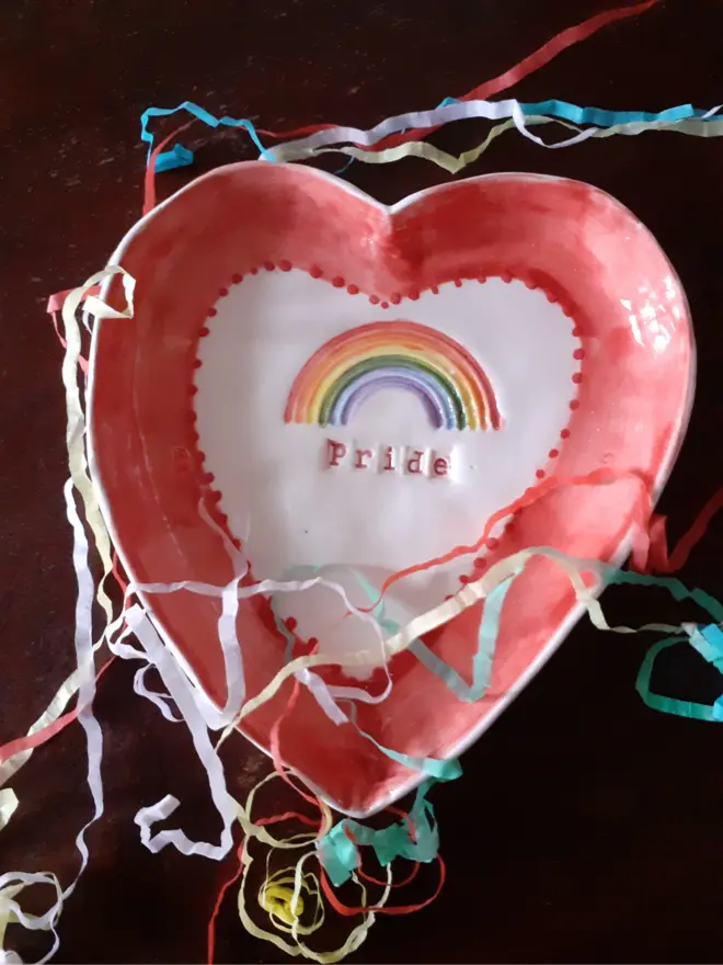 A ceramic heart shaped dish decorated with a rainbow motif and the word Pride. It has a red border.