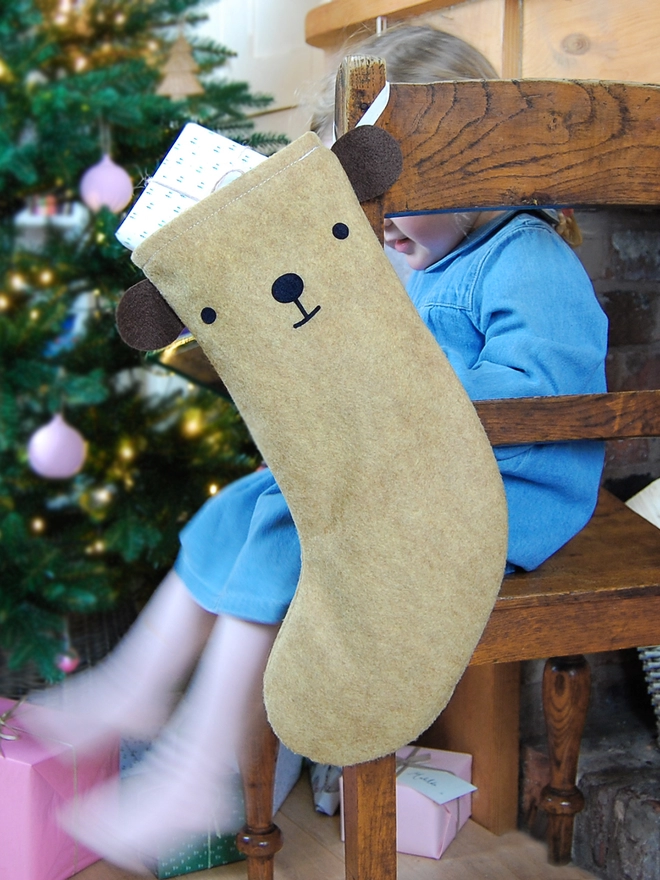 A handmade felt bear stocking hangs on a wooden chair, where a little girl sits, in front of a Christmas Tree.