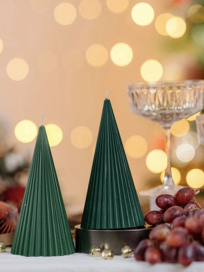 Green Christmas Tree Cone Candles. Two forest green ribbed cone candles, one raised on a dark stand, beside crystal coupe glasses and red grapes on a linen tablecloth. The fine vertical ridges give these minimalist tree candles an elegant, festive look.