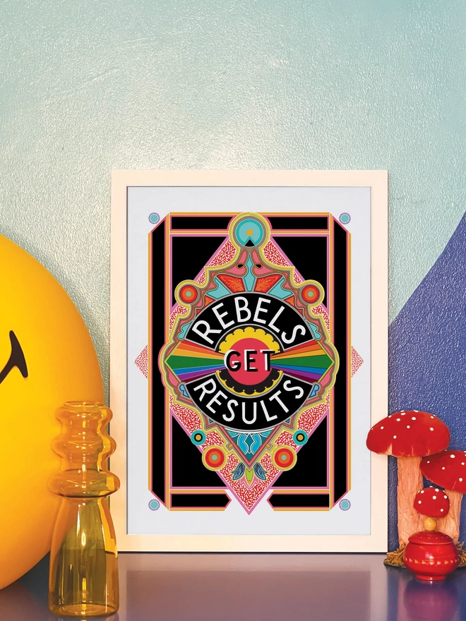 Rebels Get Results is written in white on a black background at the centre of this vibrant, abstract portrait illustration, with a black background and rainbows emitting from the centre, and multi-coloured detailing. The picture is in a white frame leaning against a painted blue wall on a blue cabinet. Next to the frame is a large illuminated yellow Smiley lamp, a yellow vase, a model of three red and white toadstools and a small round red wooden pot.