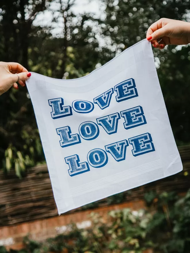 A Mr.PS LOVE LOVE LOVE hankie held aloft in a sunny garden by white hands