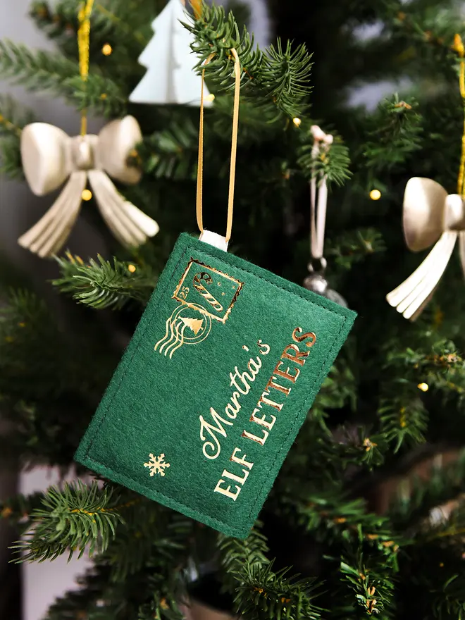 A green felt personalised envelope decoration with golden words reading "Martha's Elf Letters" and a candy cane design stamp in the corner, hangs on a decorated Christmas tree.