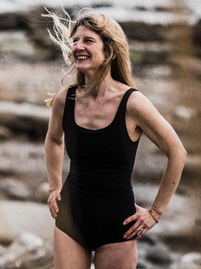 A blonde lady stood in a powerful stance with hands on hips and wind blowing her blonde hair. Wearing a Davy J Sustainable Waterwear cropped black swim top with square neck and shoulder straps and a pair of black high waist briefs