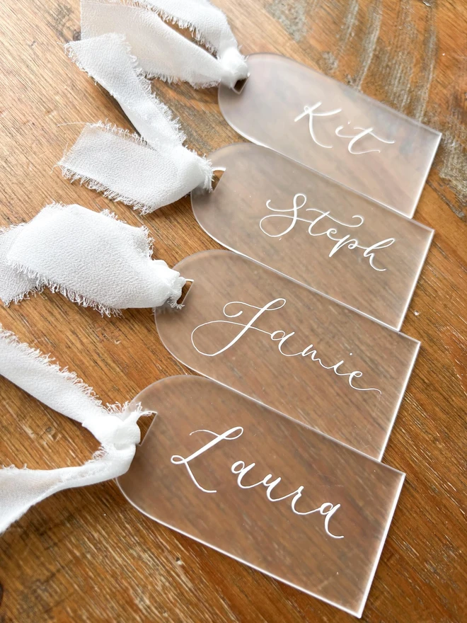 Personalised Calligraphy Frosted Acrylic Place Cards, frosted acrylic place cards with white chiffon ribbons grouped together on a wooden table. 