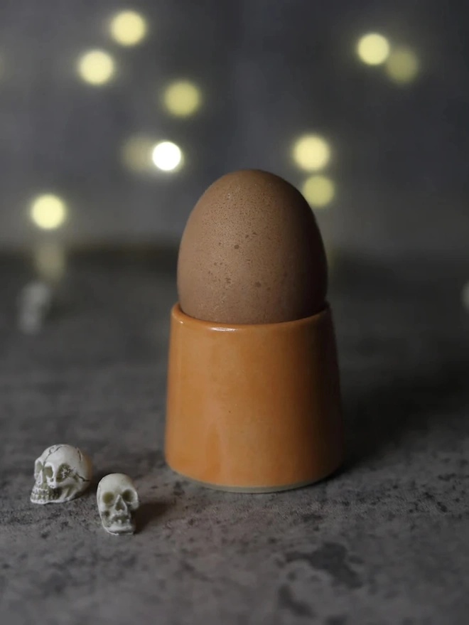 Classic Handmade Egg Cup, a ceramic egg cup sitting on a dark surface board against a fairy light backdrop. It is accompanied by some mini skulls. 