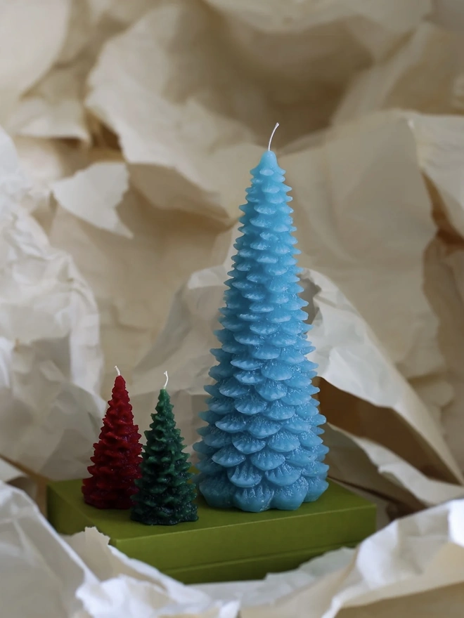 Large Christmas Tree Candle. a large sky blue christmas tree shaped candle stands on a green box, nestled among crumpled cream tissue paper. a smaller red and a smaller dark green tree candle sit in front of it for scale, both with visible wicks. the textured, layered pine branch design and slightly variegated wax give all three candles a realistic, hand-poured finish.