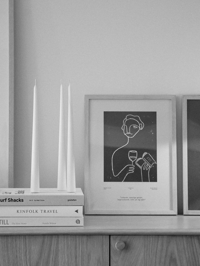 White Stem Candles (Set Of 3). A black and white image of three tall cream taper candles standing freely on a stack of coffee table books, beside a framed line-art print on a wooden sideboard. The slim freestanding design suits minimalist, gallery-style interiors.
