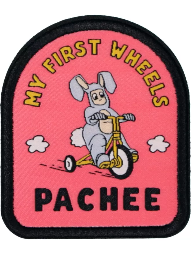 A pink patch with my first wheels sewn in yellow lettering at the top. In the centre is a kid dressed in a rabbit costume riding a bicycle with plumes of smoke emanating from the wheels. 