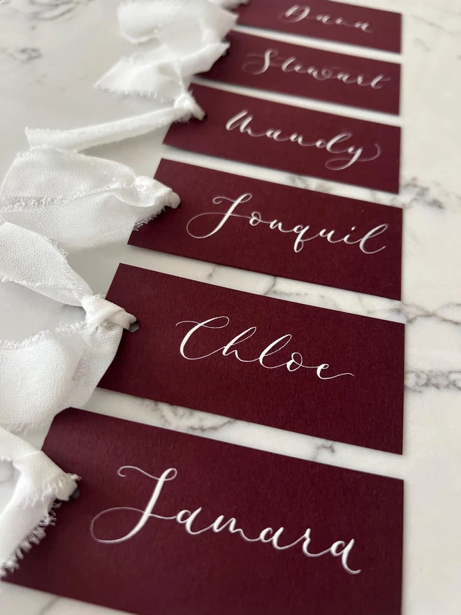 Personalised Calligraphy Wedding Burgundy Place Cards, burgundy place cards set together on a wooden table with white ribbons. 