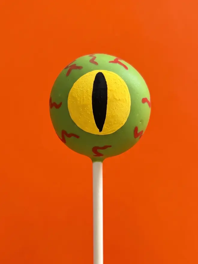 Green and yellow monster eyeball cake pop on a white stick against a bright orange background