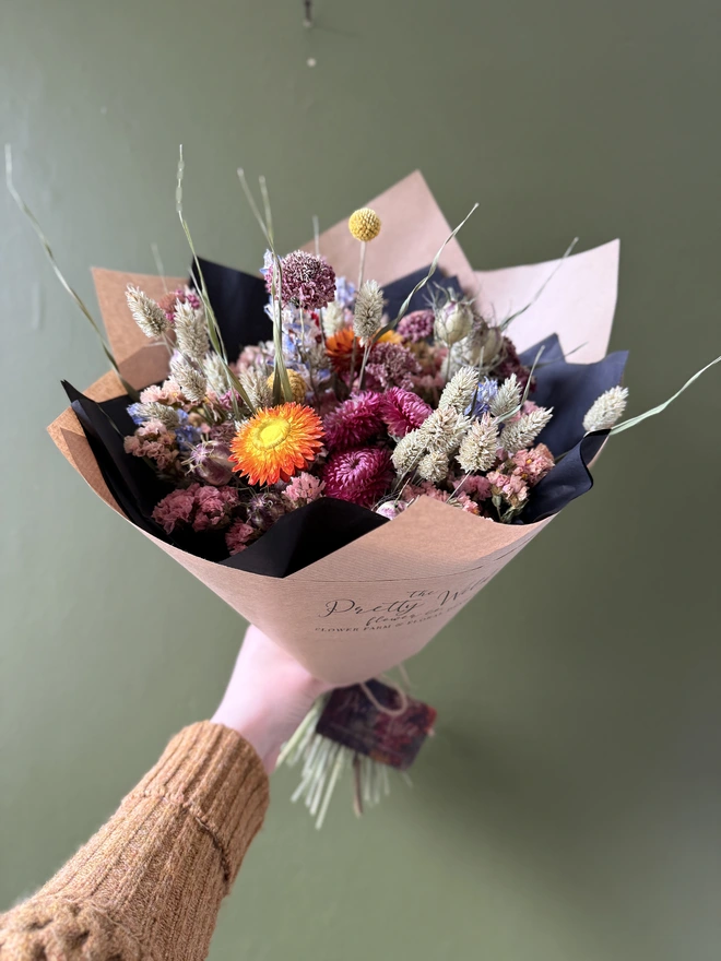 a dried flower bouquet in bright colours wrapped in black tissue and kraft paper held by a person in a mustard coloured jumper with their left arm against a green wall