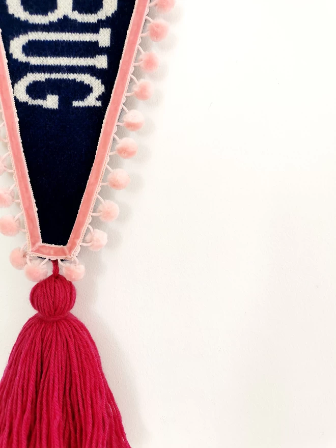 A close up product image of the front of a navy knitted pennant flag wall hanging. The end of the word ‘Bah Humbug’ can be knitted in white lambswool with a hint of gold sparkle in the text.You can see the letters ‘BUG’ here in detail alongside the blush pink pom pom trim, raspberry pink oversized tassel.