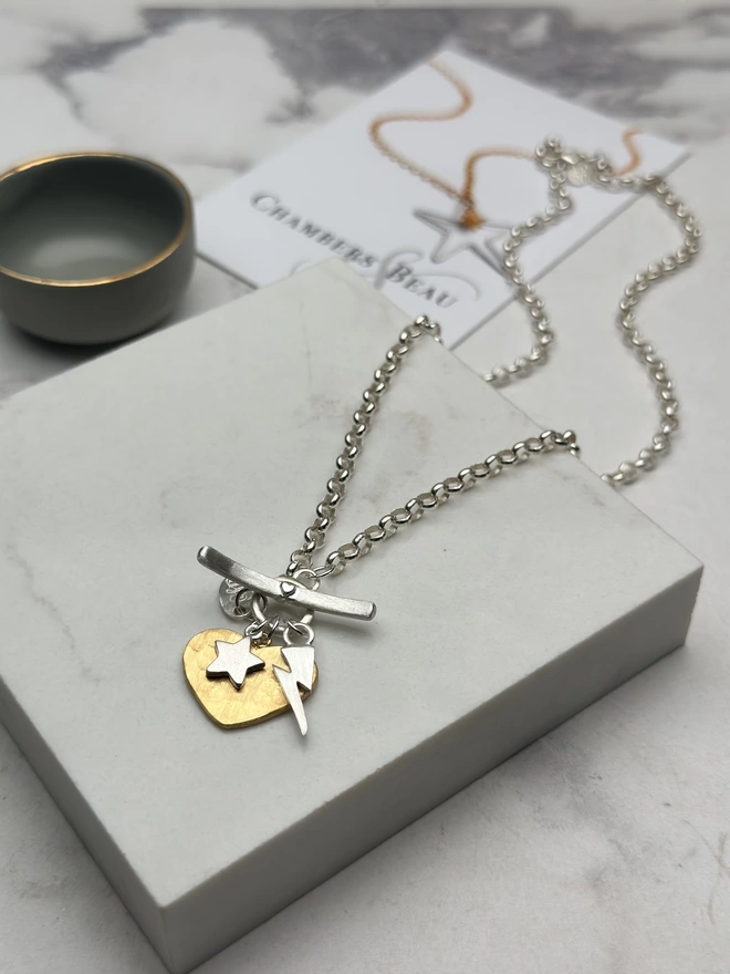 Large hammered gold heart and small silver star and electric bolt hanging from sterling silver chain with silver T-bar fastener