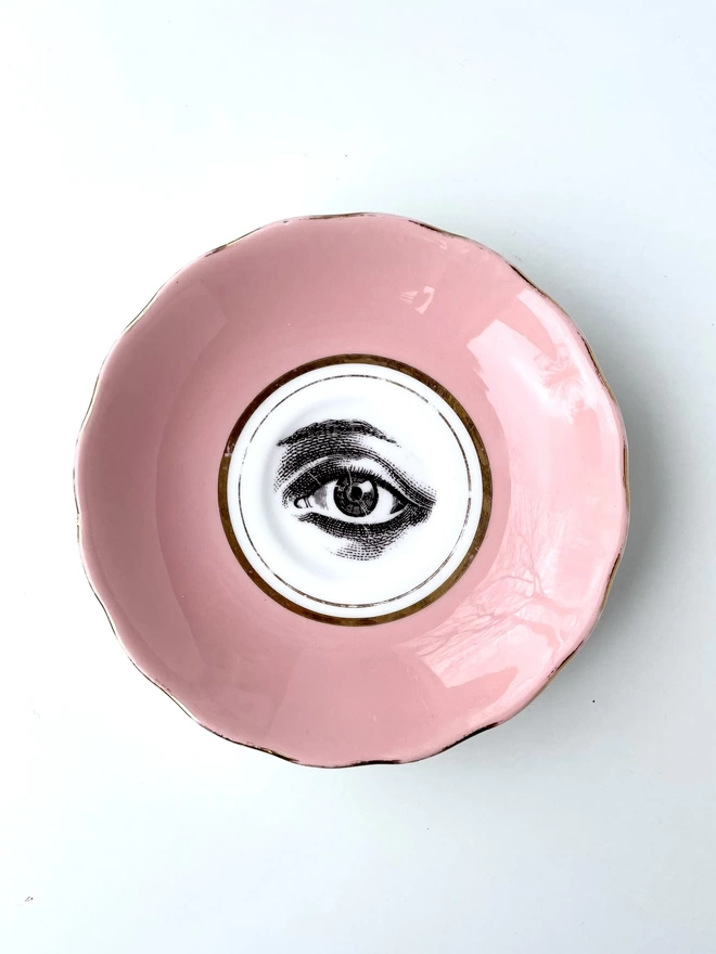 vintage pastel pink plate with a gold trim border, with a printed vintage illustration of an eye in the middle 