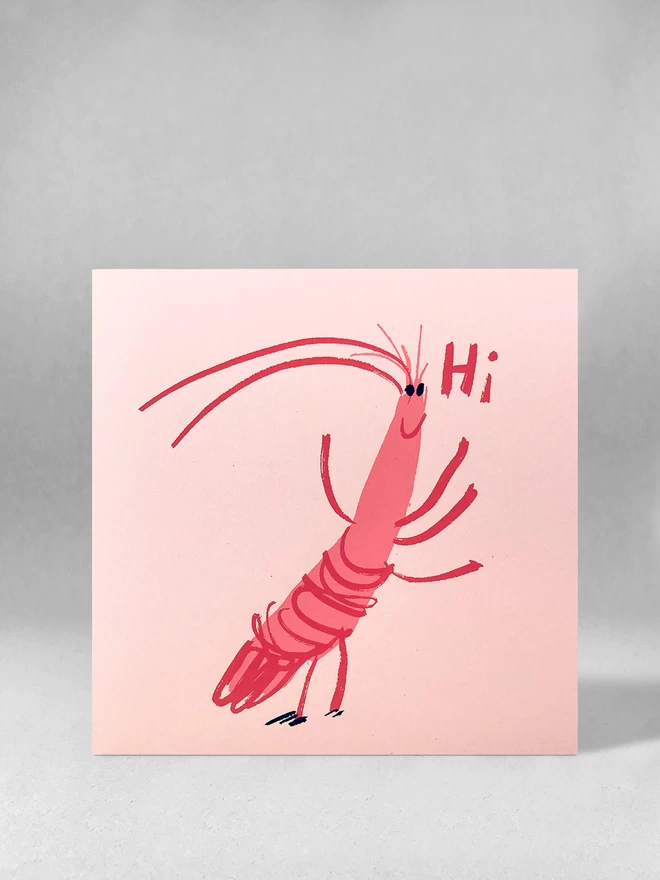 A happy cartoon prawn says Hi on this jolly upbeat pink card, printed in two pink inks with black eyes. Card is angled face on, and it is stood on a light grey background. 