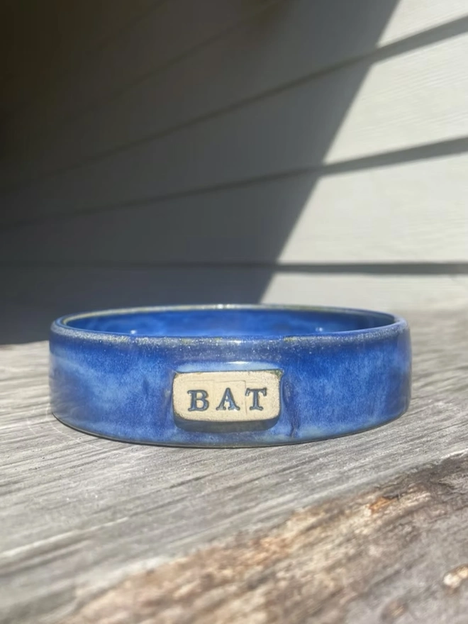 Personalised Handmade Ceramic Cat And Dog Bowls, blue pet bowl that says 'Bat' on a wooden surface. 