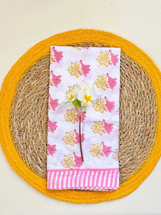 Close up of a round woven seagrass placemats with yellow rings, shown on a light surface with small white flowers.